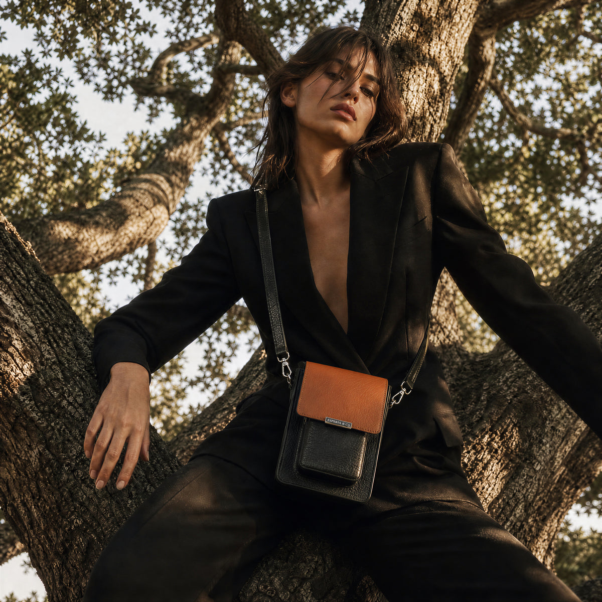 Model in a black outfit with a brown leather crossbody bag sitting in a tree